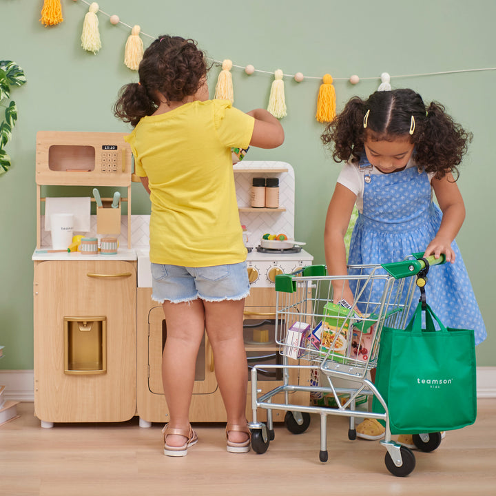 Two children playing with a Teamson Kids - Little Helper Dallas Shopping Cart with Play Food, Chrome/Green and toy kitchen.