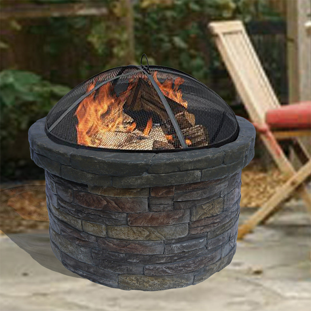 Teamson Home 27" Outdoor Round Stone Wood Burning Fire Pit with Steel Base, Natural Stone with blazing fire in a backyard setting.