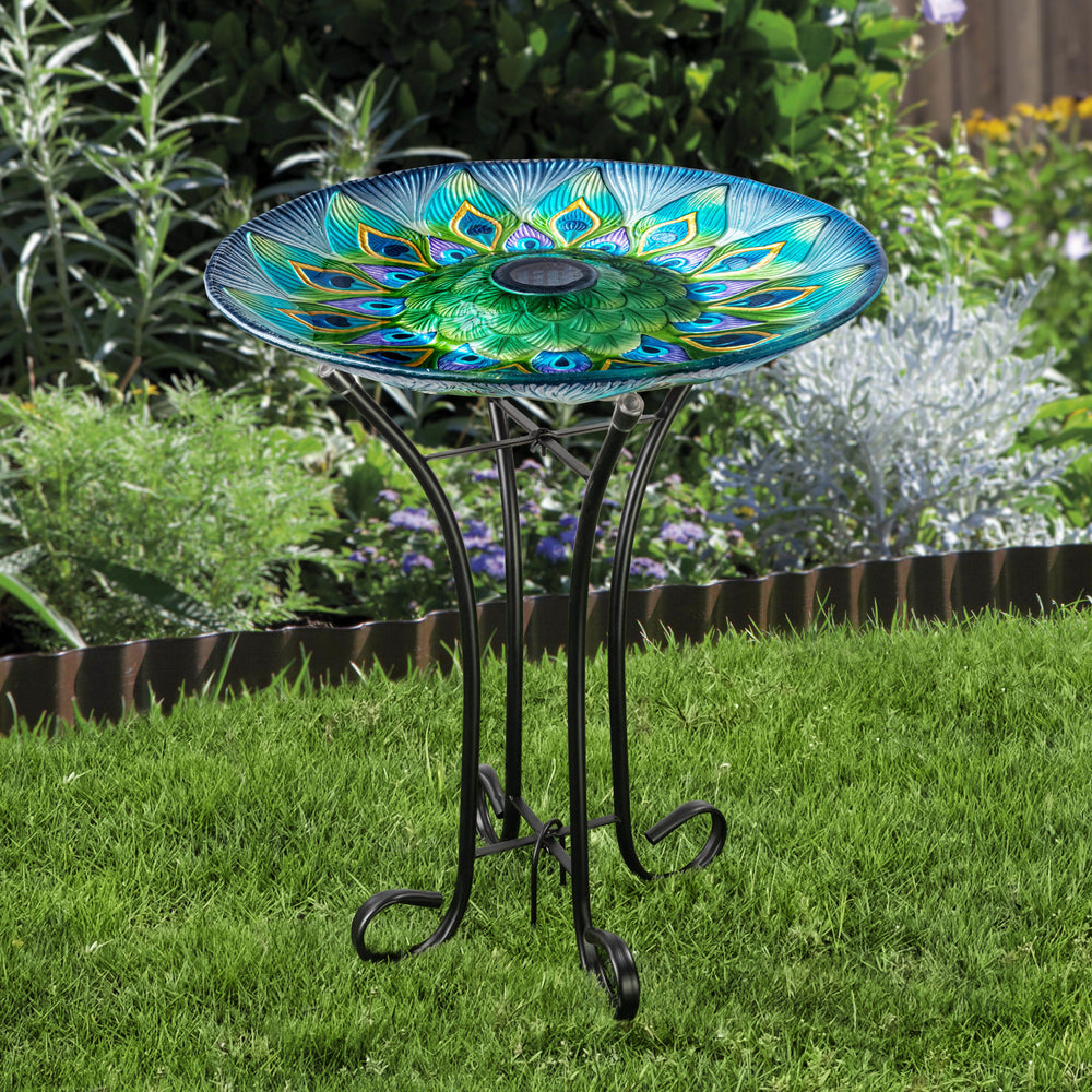 A 17.8" Peacock Fusion Glass Birdbath with Solar-Powered Light, Blue and Green, on a metal stand in a garden setting.