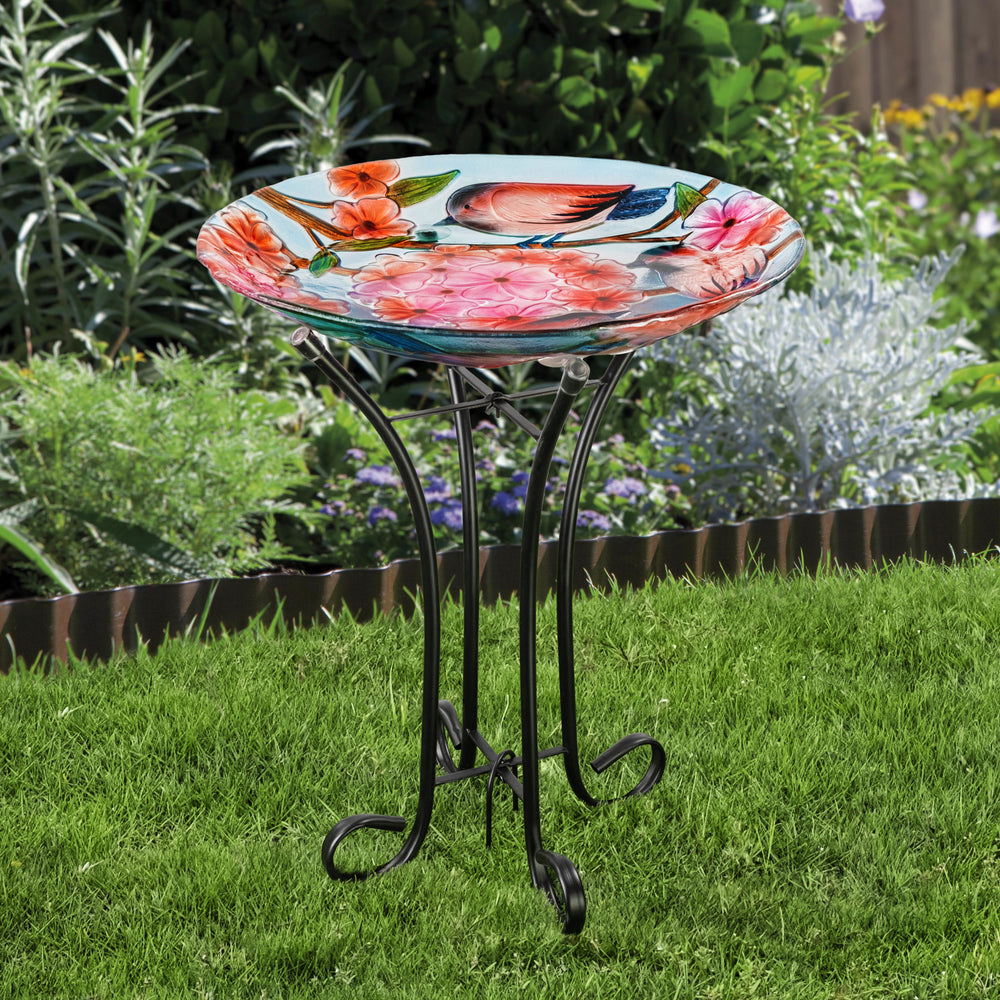 A 17.8" Robins & Blossoms Fusion Glass Birdbath with Metal Stand, Multicolored, with flowers on it in a garden.