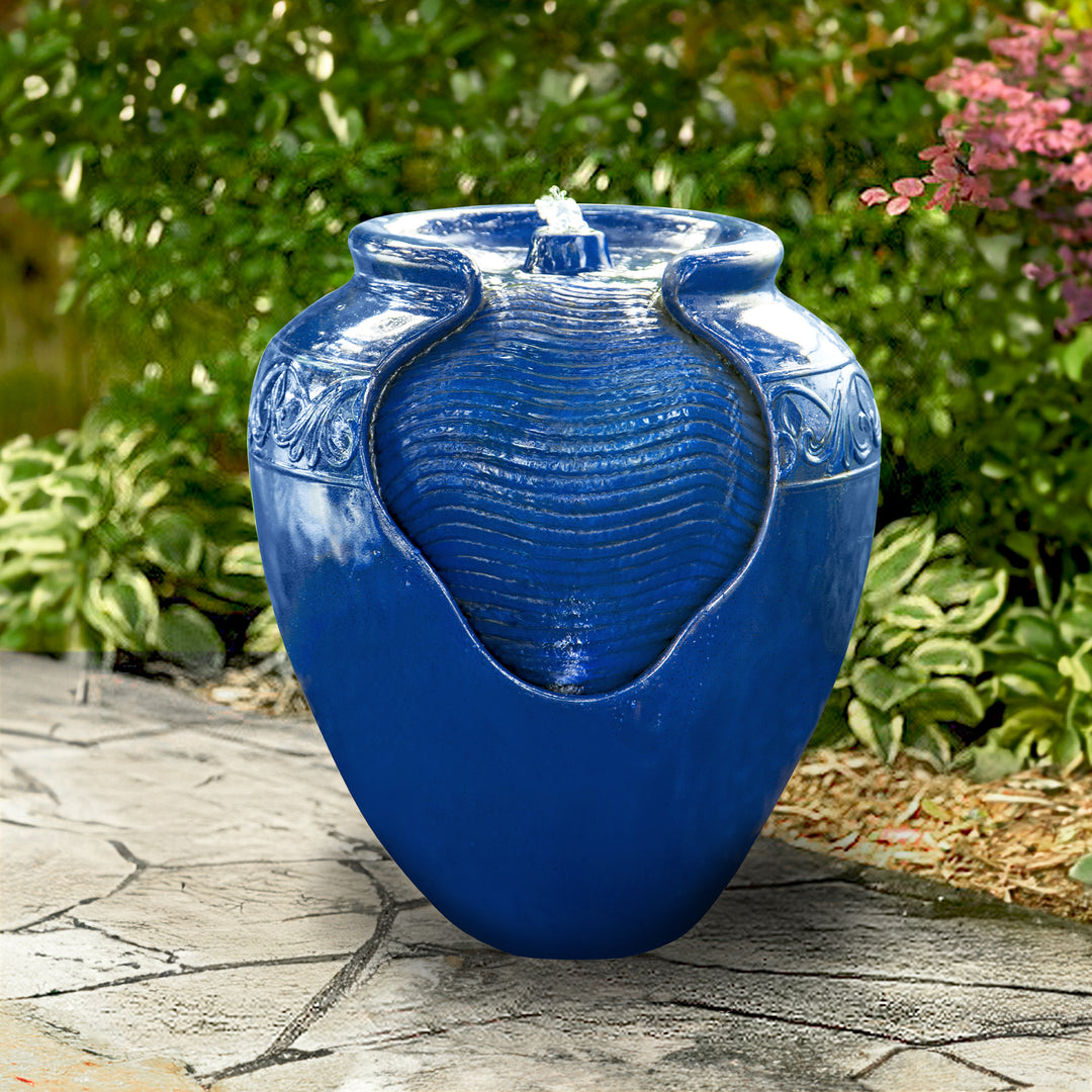 A Teamson Home Outdoor Glazed Pot Floor Fountain with LED Lights, Royal Blue, in the shape of a jar, located outdoors on a stone path surrounded by greenery.