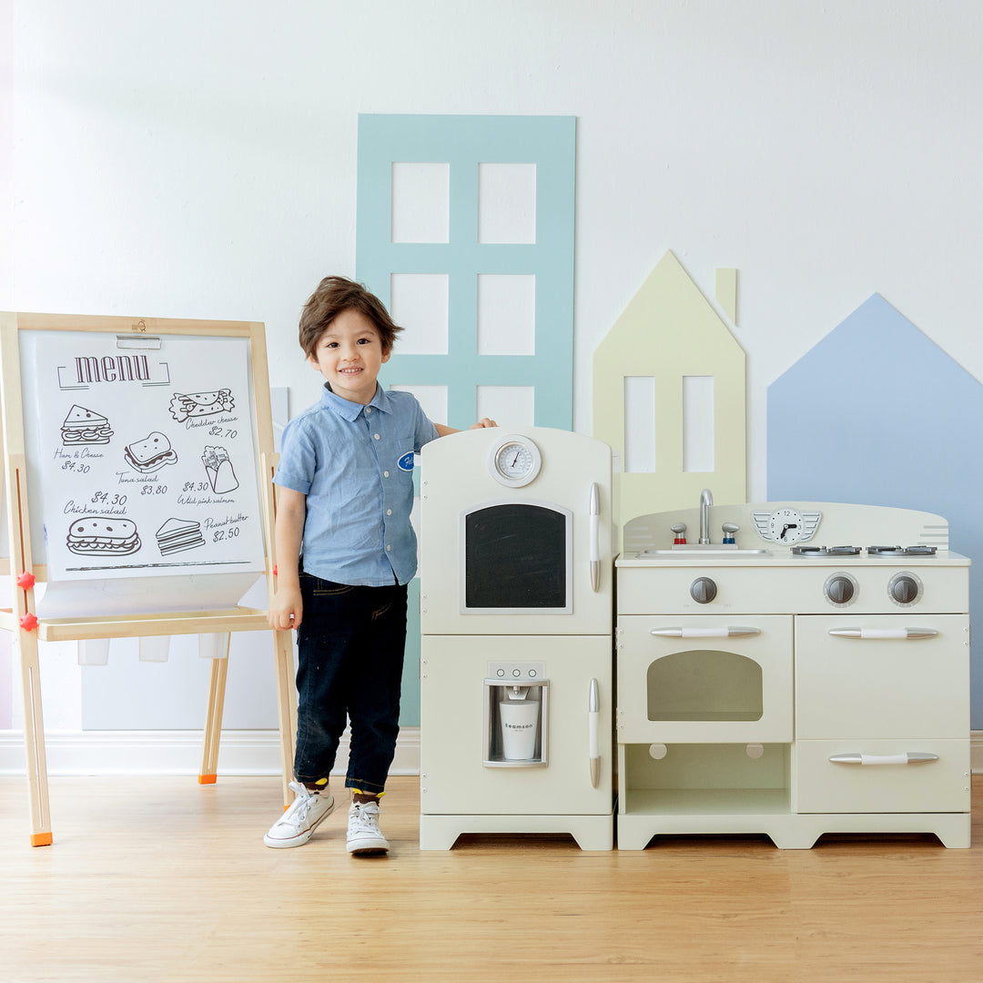A child standing beside a Teamson Kids Little Chef Fairfield Retro Kids Kitchen Playset with Refrigerator, Ivory and a menu board in the background.