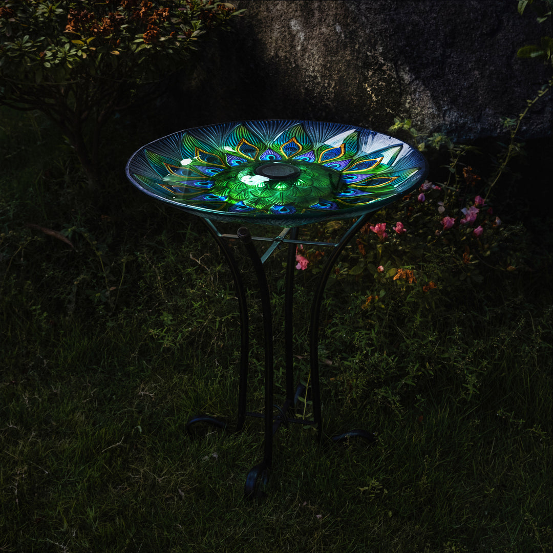 A 17.8" Peacock Fusion Glass Birdbath with Solar-Powered Light, Blue and Green, illuminated, on a metal stand in a garden at night.
