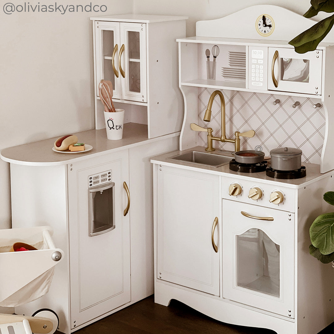 Modular play kitchen in white with gold and gray accents with cabinets, microwave, sink, ice maker, oven and stovetop. with pretend food on a plate.
