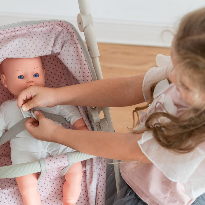 A little girl buckling her baby doll into a pink with gray polka dots convertible doll stroller.