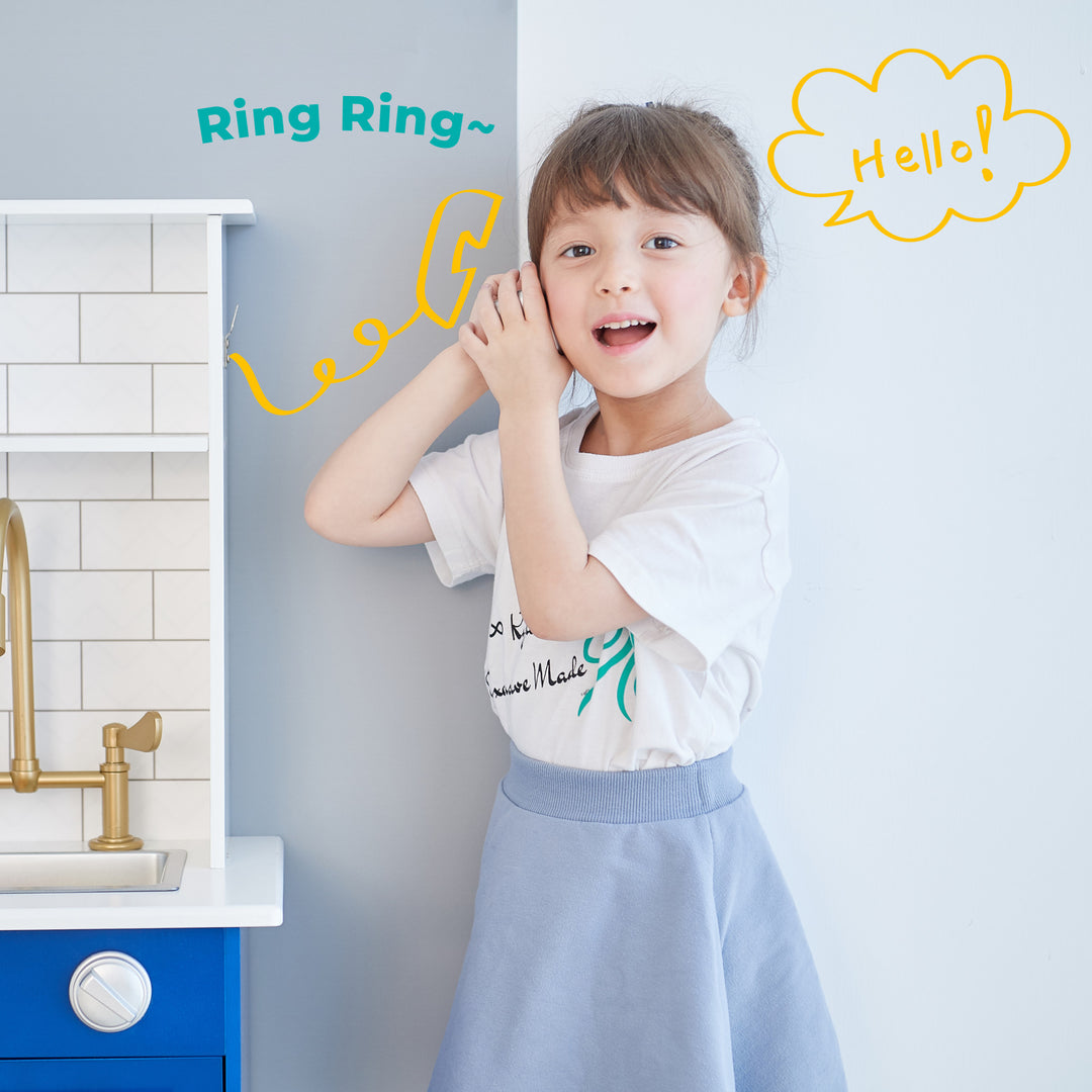 A young girl pretending to talk on the phone with hand gestures and illustrated sound effects next to her Teamson Kids Little Chef Berlin Play Kitchen with Cookware Accessories, White/Blue.