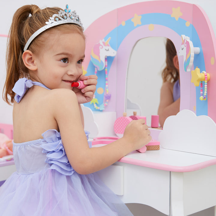 A little girl putting on pretend lipstick sitting at a white vanity table and stool with a rainbow, unicorns, stars, and a mirror.