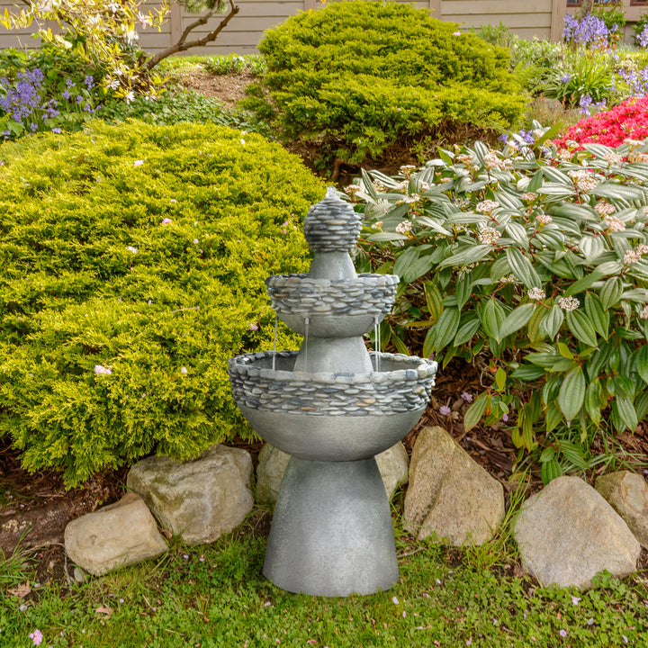 A Teamson Home Outdoor 3-Tier Pedestal Floor Fountain, Gray, set next to some bushes and flowering plants outside a home