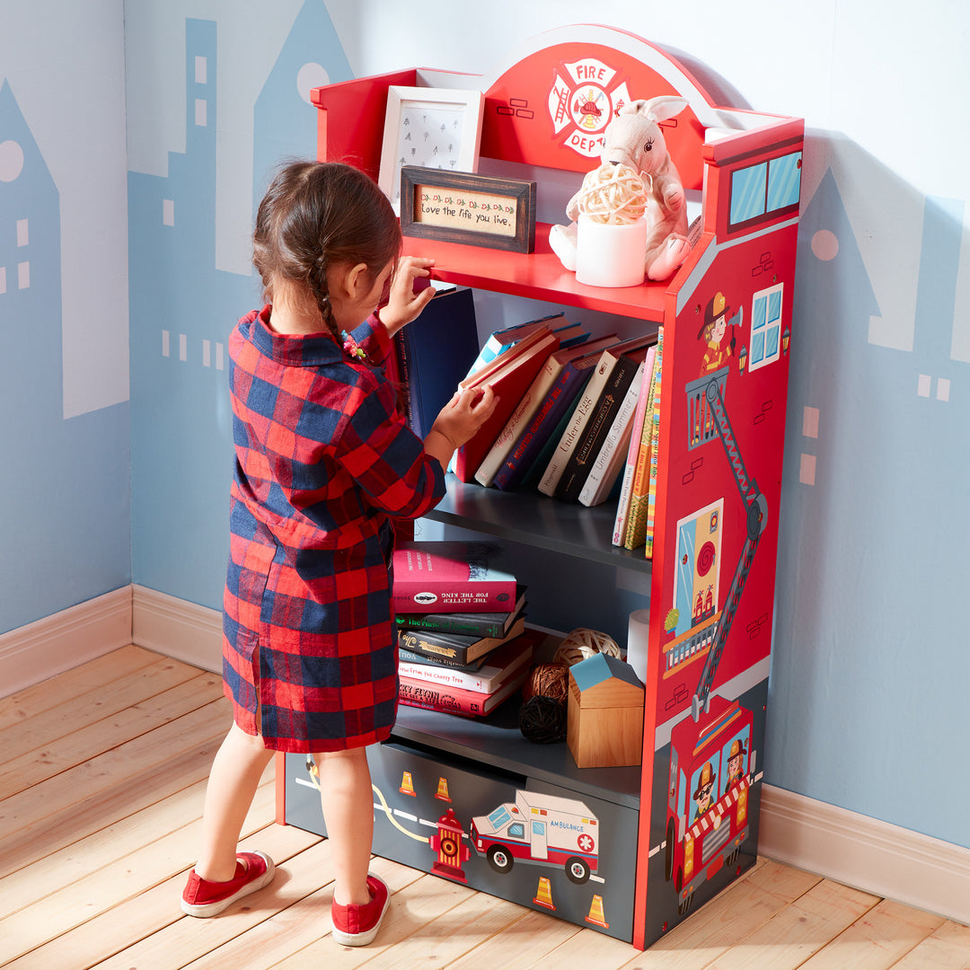 A little girl standing in front of a Fantasy Fields Little Fire Fighters Bookshelf with Drawer, Red.