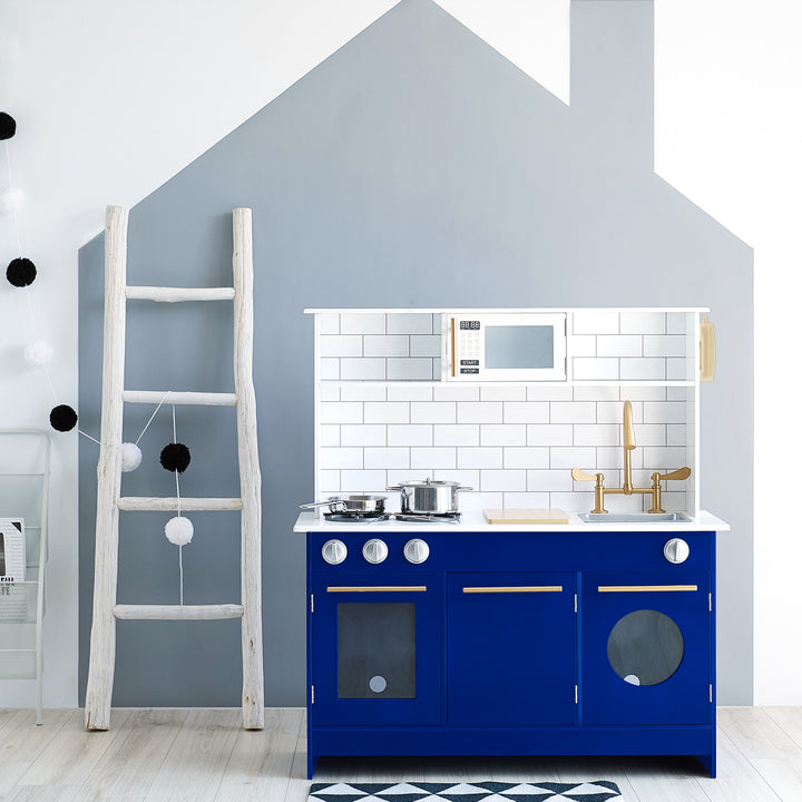 A stylish children's Teamson Kids Little Chef Berlin Play Kitchen with Cookware Accessories, White/Blue, with a blue cabinet, white backsplash, and wooden accents against a wall with a whimsical house-shaped paint design.