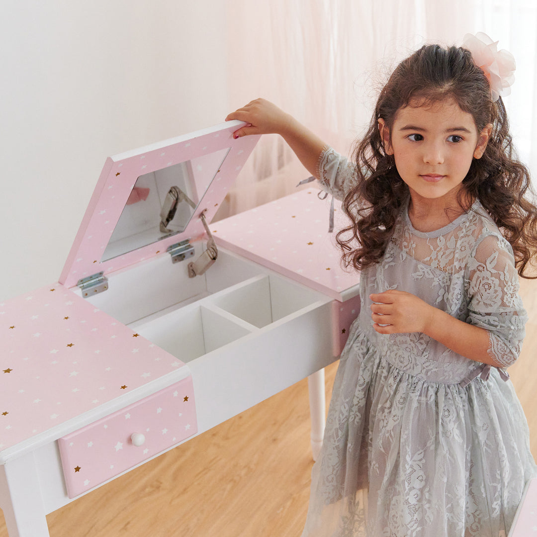 A little girl standing next to a Fantasy Fields Kids Kate Twinkle Star Vanity Set with Foldable Mirror and Chair by Teamson Kids.