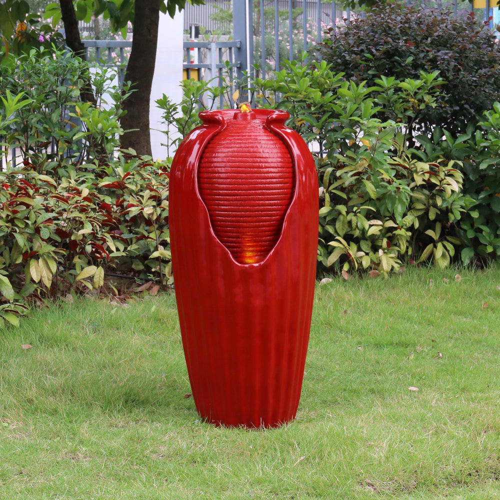 A large red Teamson Home Indoor/Outdoor Contemporary Glazed Contoured Vase Water Fountain outdoors surrounded by plants and illuminated by LED lights.
