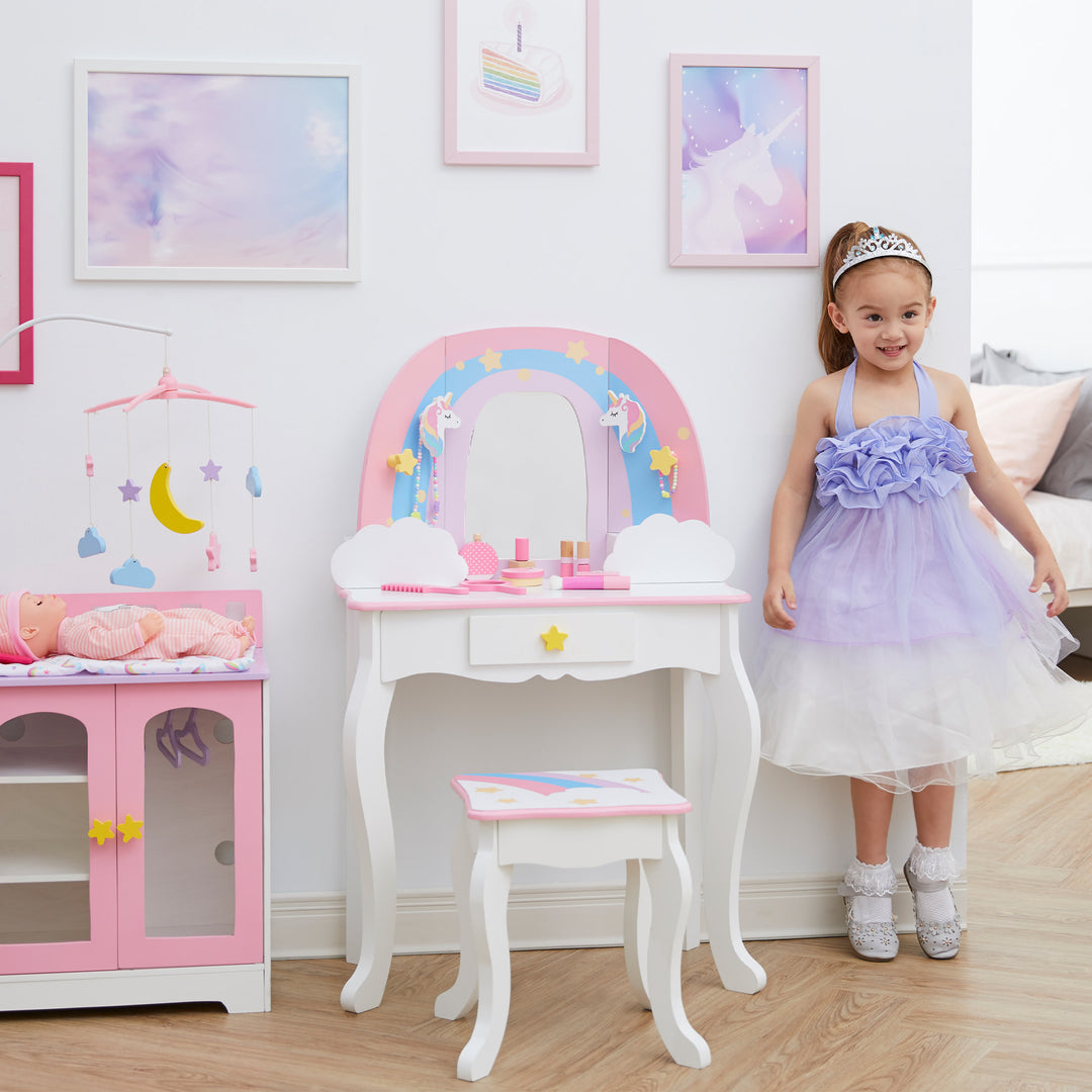 A little girl in a lavender dress standing next to a white vanity table and stool with a rainbow, unicorns, stars, and a mirror.