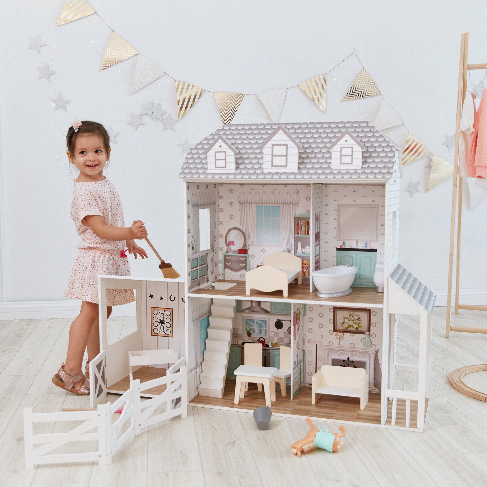 Toddler girl standing in front of a large doll house. Doll House has multiple rooms with accessories and a barn.