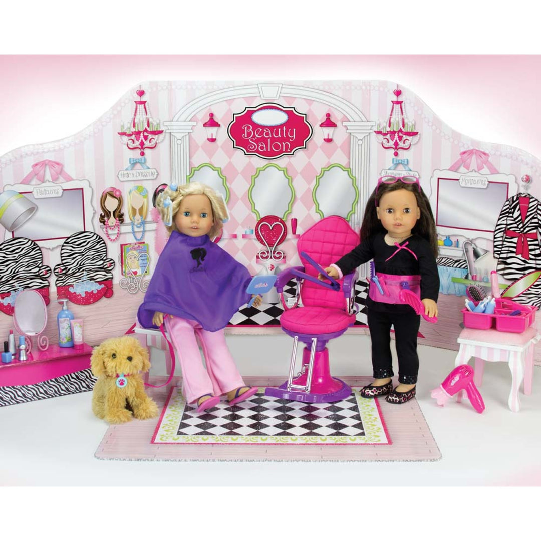 A blonde 18" doll in a purple smock standing next to a salon chair. A brunette doll dressed as a hair dresser in black stands on the other side of the chair.