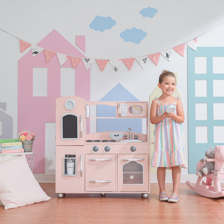 A young girl smiling in a pastel-colored playroom with a Teamson Kids Little Chef Westchester Retro Play Kitchen, Pink that includes interactive features like a toy telephone.