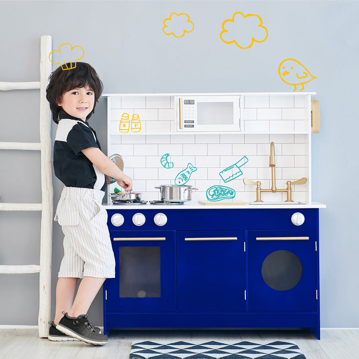 Child pretending to cook in a Teamson Kids Little Chef Berlin Play Kitchen with Cookware Accessories, White/Blue playset with playful wall doodles.