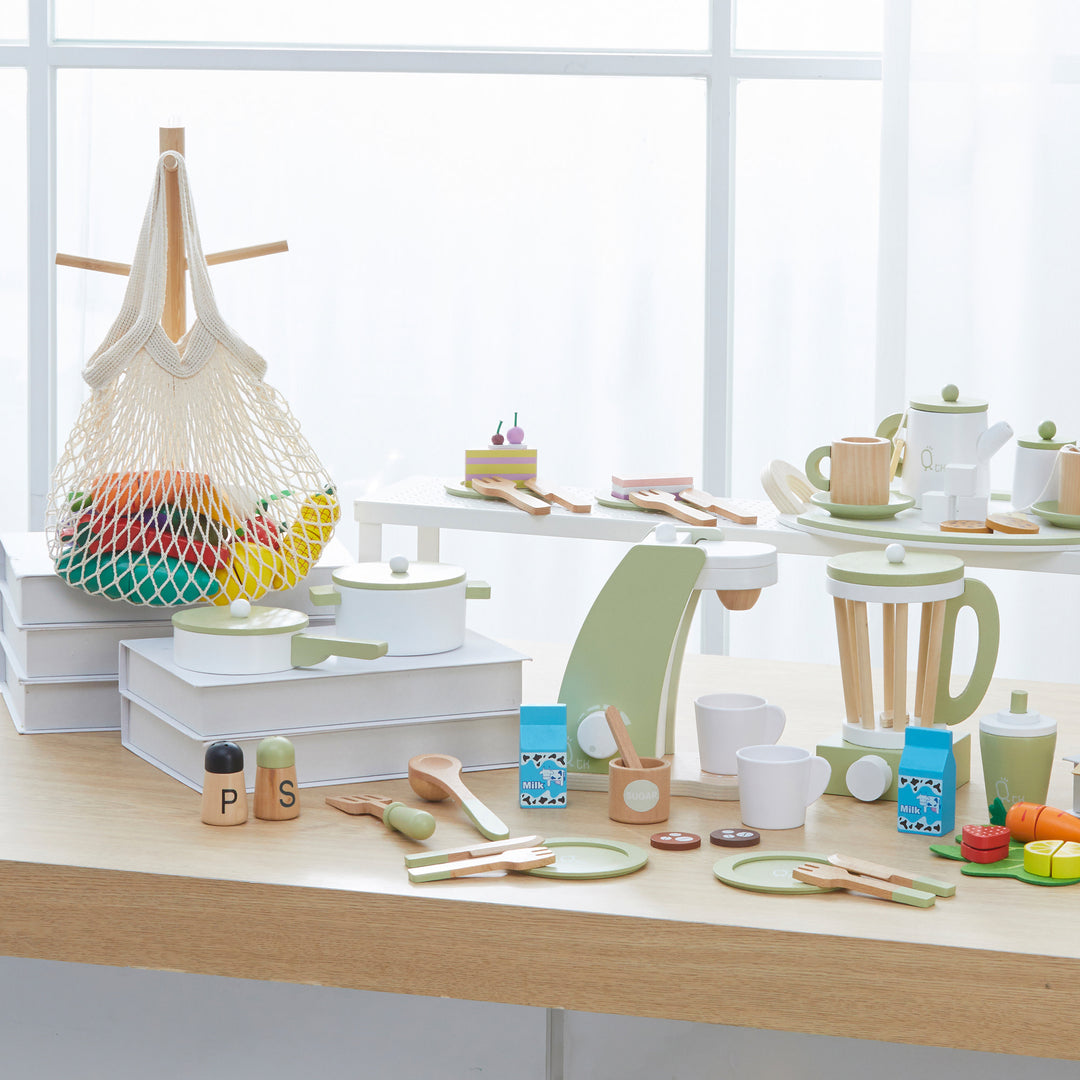 A modern kitchen table set with various Teamson Kids Little Chef Frankfurt wooden cookware play kitchen accessories, dishes, and a fruit basket in a bright, airy room, featuring kid-friendly dimensions.