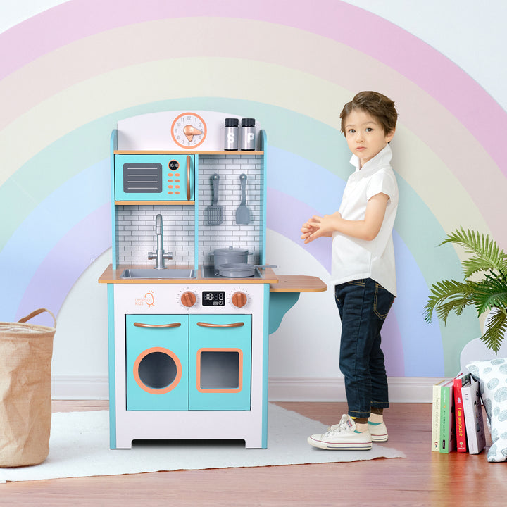 A little boy standing next to a play kitchen against a rainbow-painted wall in a playroom.