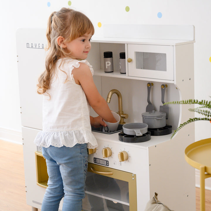 A young girl plays with a Teamson Kids Little Chef Mayfair Classic Kids Kitchen Playset with 11 Accessories, White/Gold featuring a pretend ice machine.