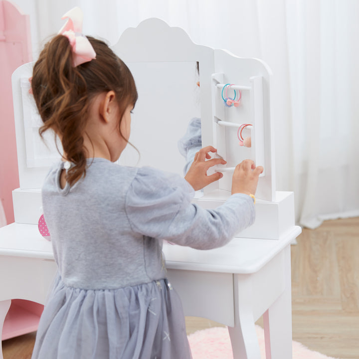 A little girl adjusting the hanging feature on her white vanity set with matching stool, storage drawers, and a mirror with storage racks on either side.