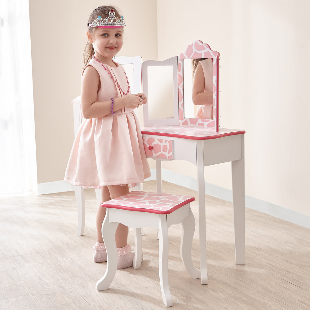 A little girl in a pink dress standing in front of a mirror in the Fantasy Fields Gisele Giraffe Prints Play Vanity Set, Pink/White.