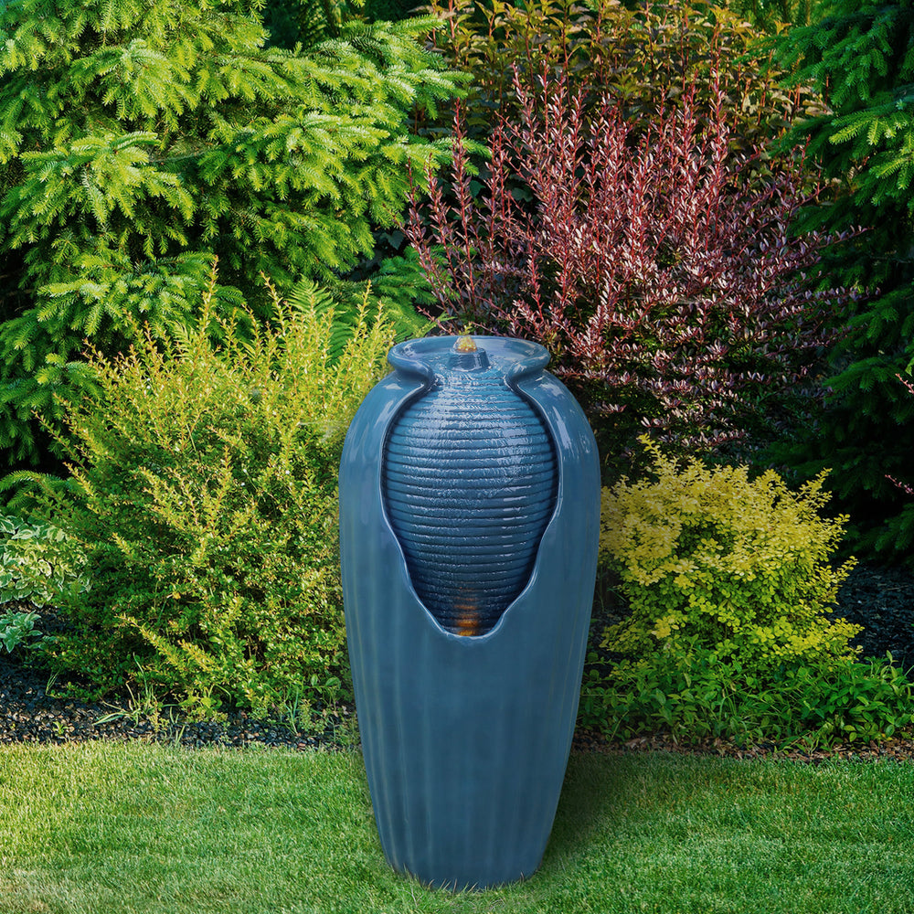 Teamson Home Indoor/Outdoor Contemporary Vase Water Fountain with LED Lights, Blue, sat against shrubs and trees in a garden.