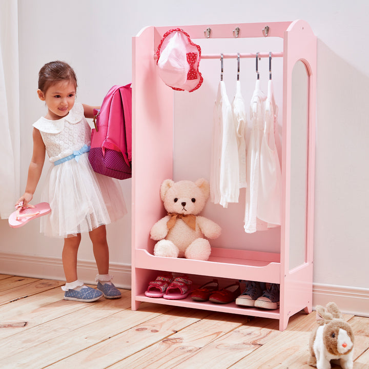 A little girl standing next to a Fantasy Fields Little Princess Clothing Rack with Storage, Pink.