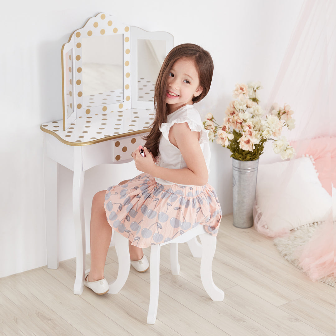 A little girl sitting on a chair in front of a Teamson Kids Gisele Polka Dot Vanity Playset, White / Gold mirror.