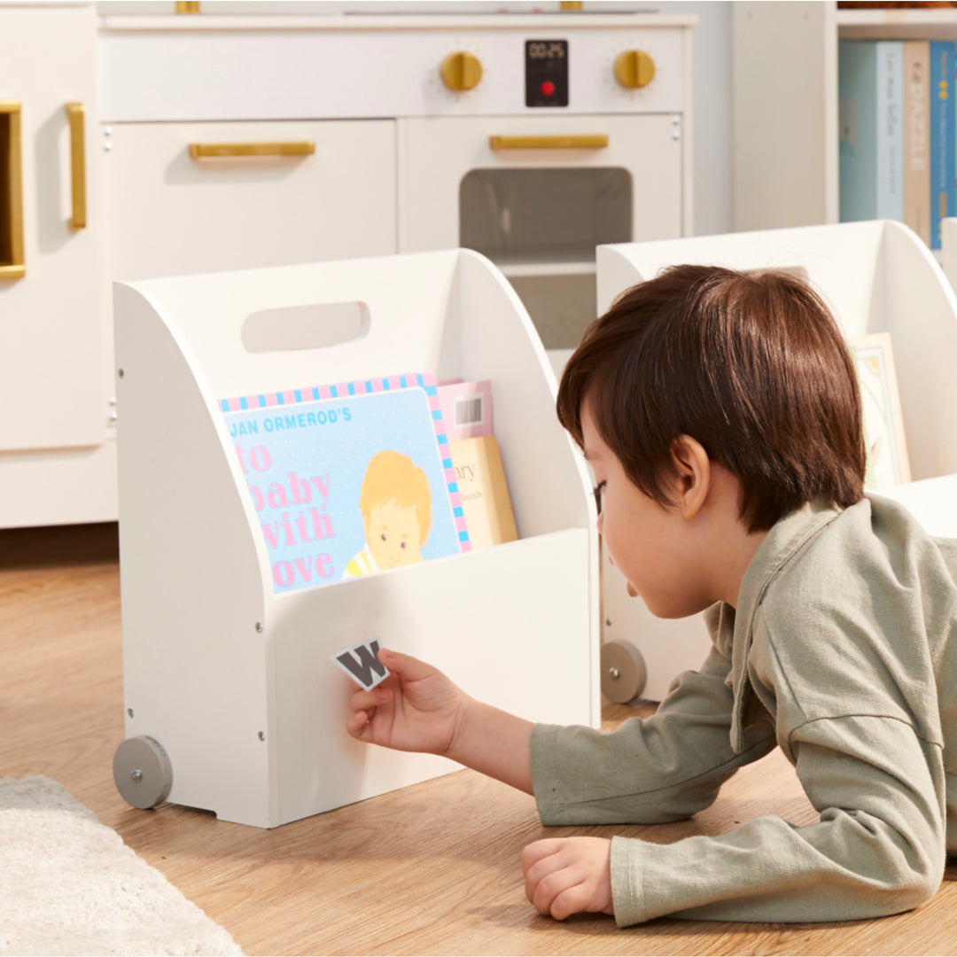 A young boy laying on the floor with a Fantasy Fields Kids Portable Bookcase Set of 3 with Magnetic Dry Erase Whiteboard, White in front of him.