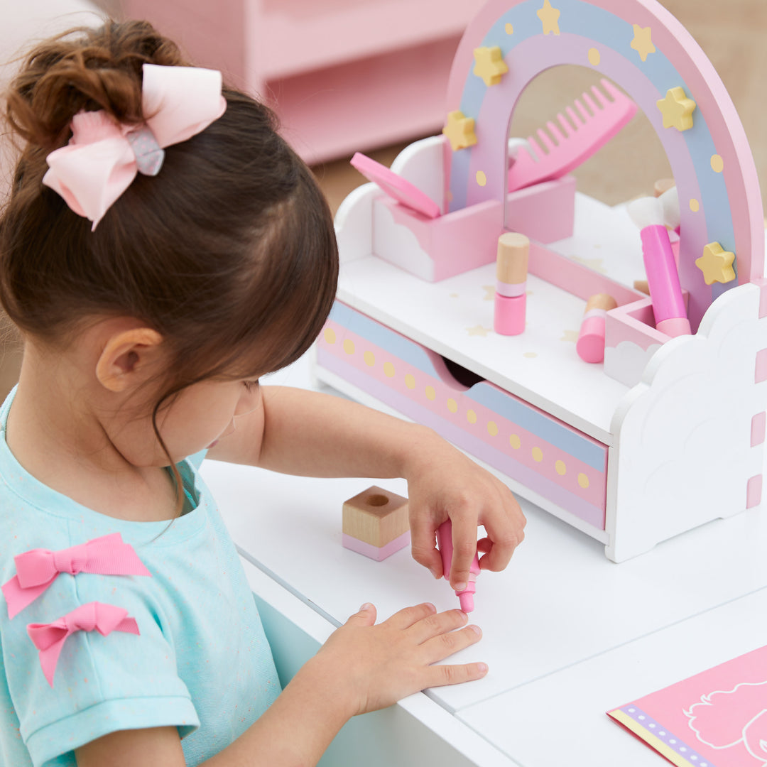 A little girl sitting next to her tabletop vanity with rainbow accents and a mirror pretending to apply pretend nailpolish.