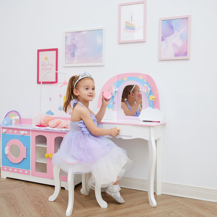 A little girl in a pink dress sitting at a white vanity table and stool with a rainbow, unicorns, stars, and a mirror.