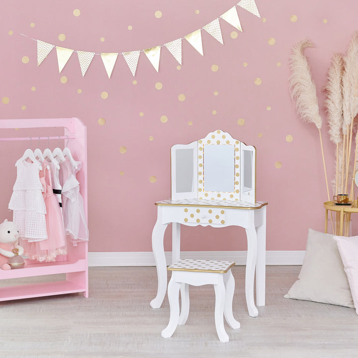 A pink bedroom with gold polka dots with a pink wardrobe with dresses hanging from hangers and a white with gold polka dots kids vanity table and stool.