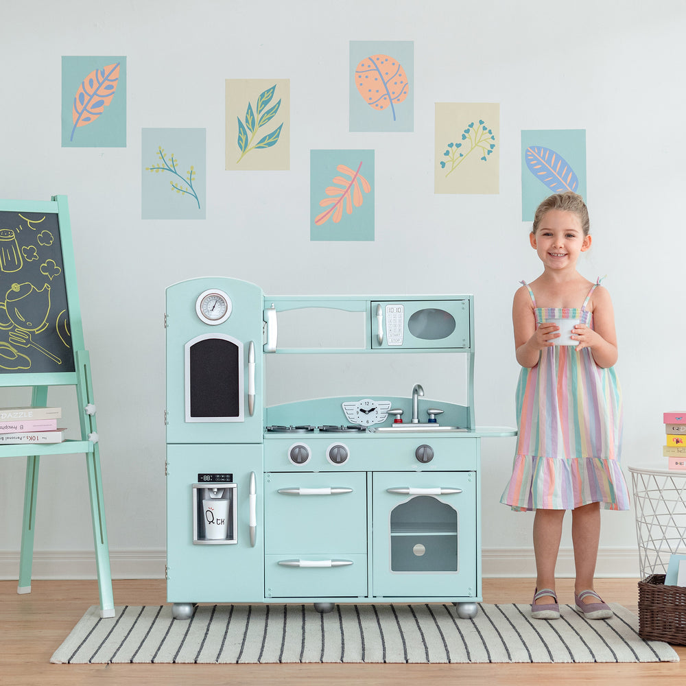 A young girl smiling and holding a cup, standing next to a Teamson Kids Little Chef Westchester Retro Kids Kitchen Playset, Mint with drawings on the wall behind her.