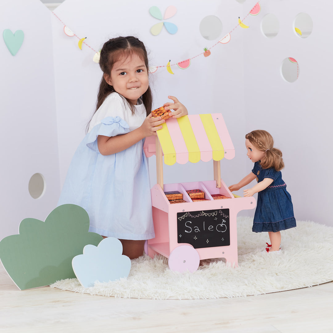 A little girl is standing next to Olivia's Little World Modern Nordic Princess Doll Pastry Cart with Fruit Boxes, Pink.