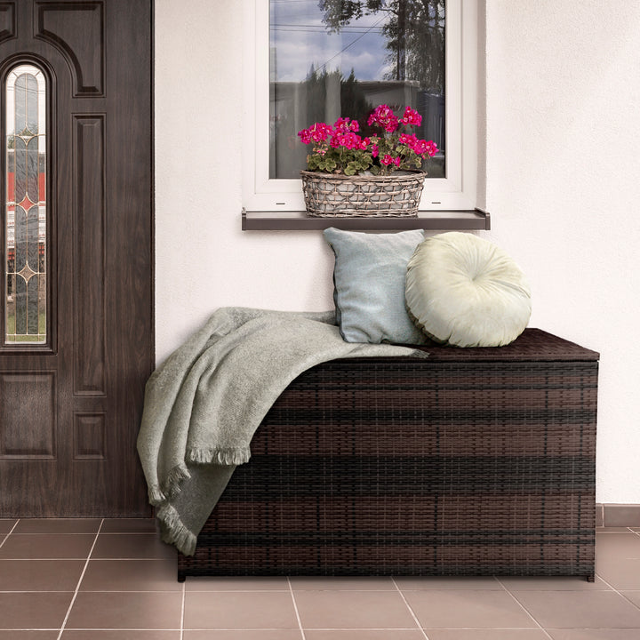 Teamson Home Brown PE Rattan 154-Gallon Outdoor Deck Box underneath a window on a porch with two pillows and a blanket on top