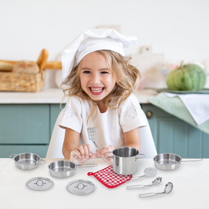 A joyful young child wearing a chef's hat and apron while playing with the Teamson Kids 11 Piece Little Chef Frankfurt Stainless Steel Cooking Accessory Set on a table.