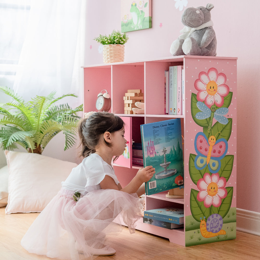 A girl in a tutu playing with a book from her Fantasy Fields Kids Painted Wooden Magic Garden Adjustable Cube Bookshelf, Pink.