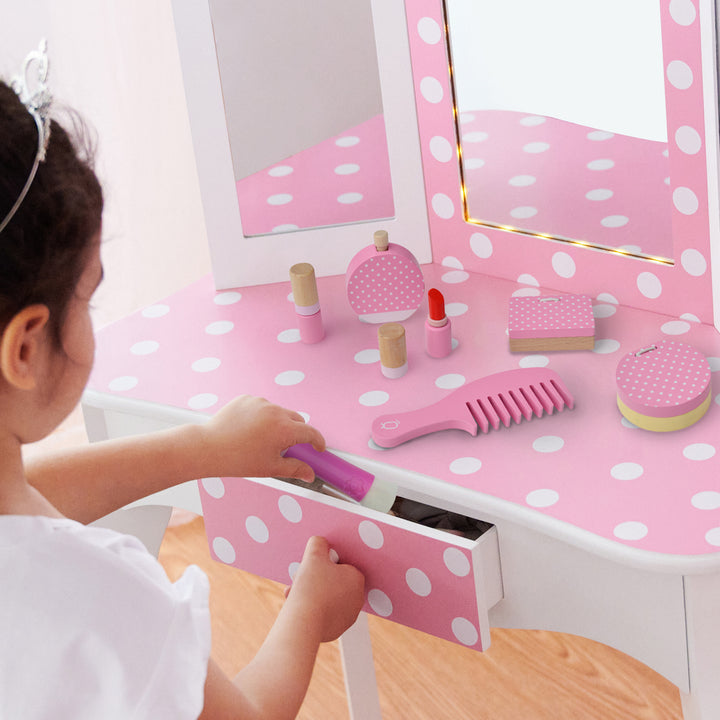 A little girl sitting at a white play vanity table with pink accents and white polka dots putting a make-up brush in a drawer. There are two lipstick tubes, an eyeshadow pallet, a compact, a comb, and perfume on the tabletop.