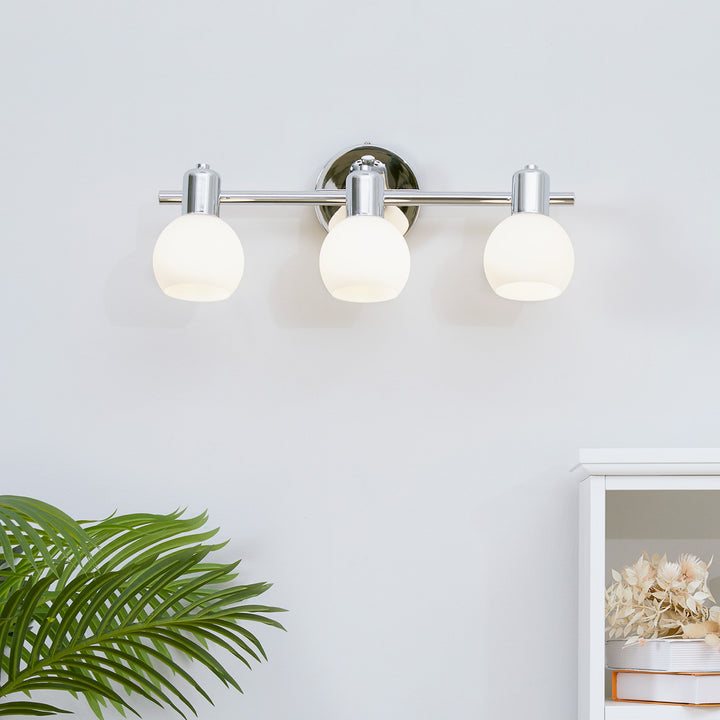 A Teamson Home  3-Light Vanity Fixture with Frosted Globe Shades, Chrome, above a palm plant and white shelving unit