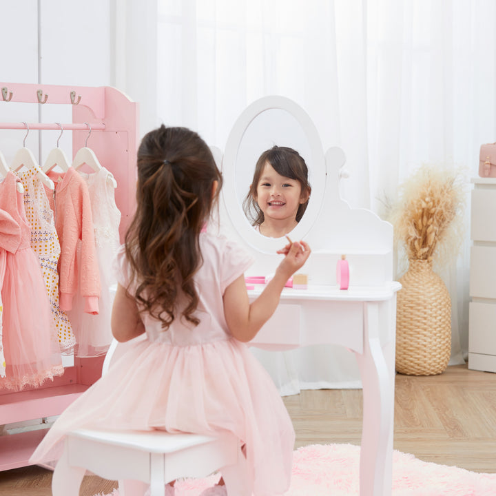 A little girl in a pink dress looking into the mirror on the white vanity set.