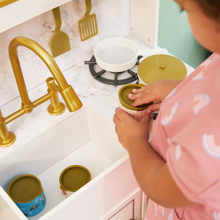 A young girl opens a pretend can of food next to the farmhouse-style sink on her white play kitchen with gold accents.