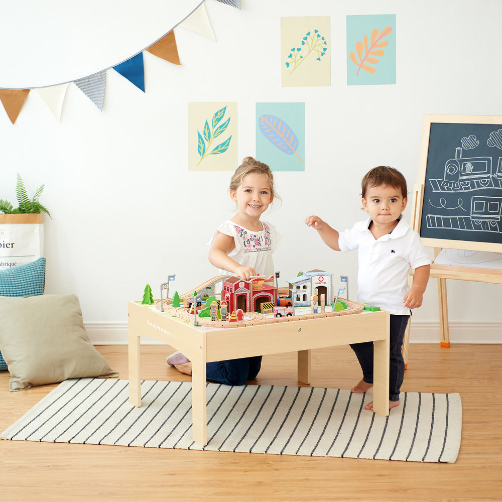 Two children playing with a Teamson Kids Preschool Play Lab Toys Wooden Table with 85-pc Train and Town Set, Natural in a brightly-lit playroom.