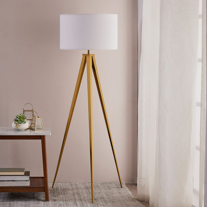 A Teamson Home Romanza 62" Postmodern Tripod Floor Lamp with Drum Shade in Matte Gold/White stands next to a window with sheer curtains and beside a small table with decorative items.