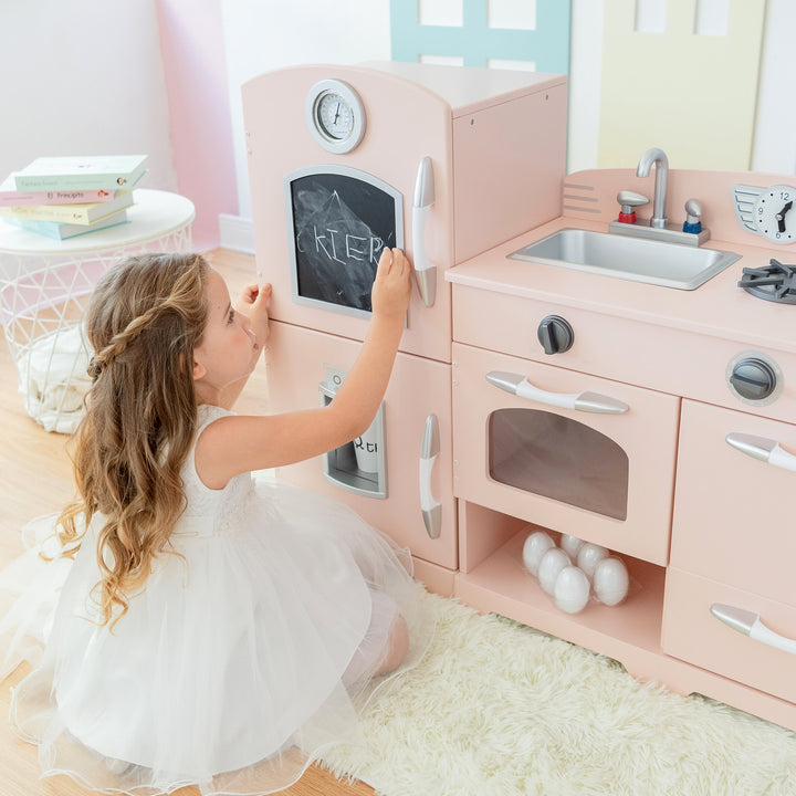 A young girl in a white dress writing on the Teamson Kids Little Chef Fairfield Retro Kids Kitchen Playset's chalkboard with interactive features.