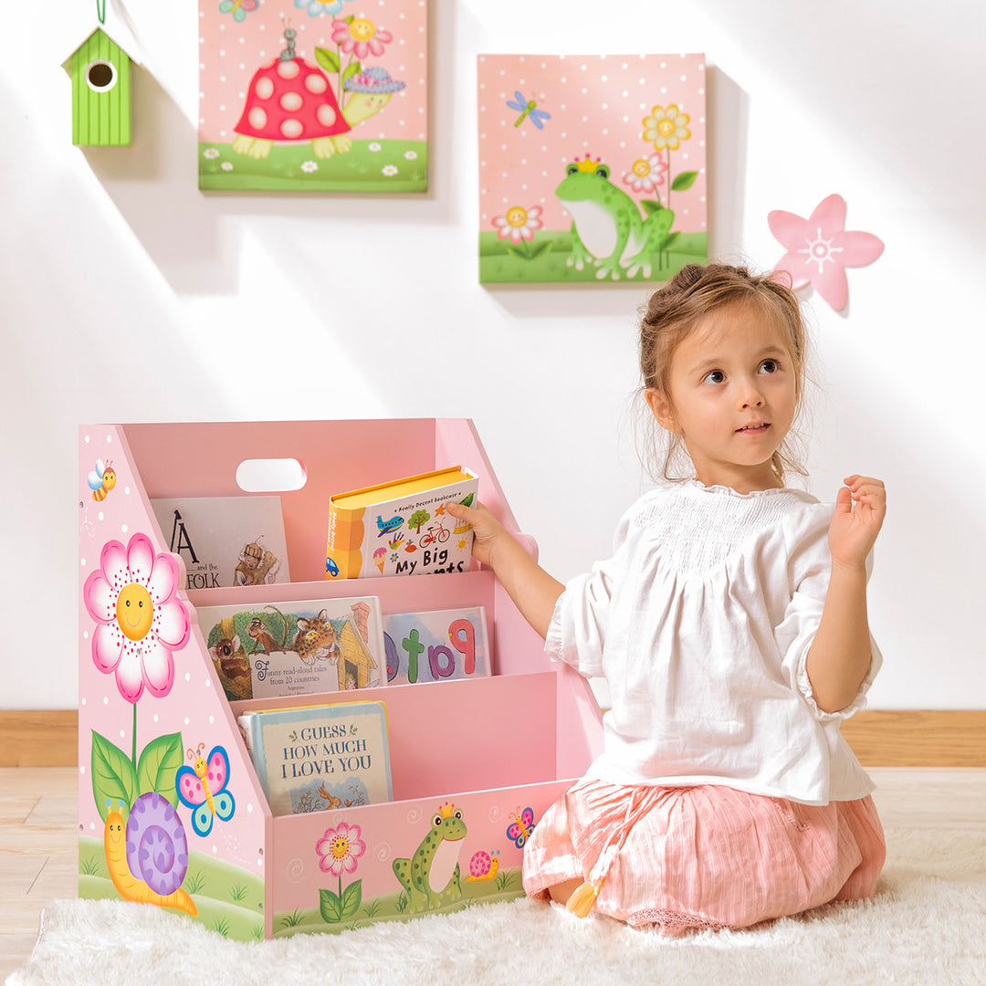 A little girl is sitting on the floor next to the Fantasy Fields Kids Painted Wooden Magic Garden 3-Tiered Bookshelf, Pink in the playroom.