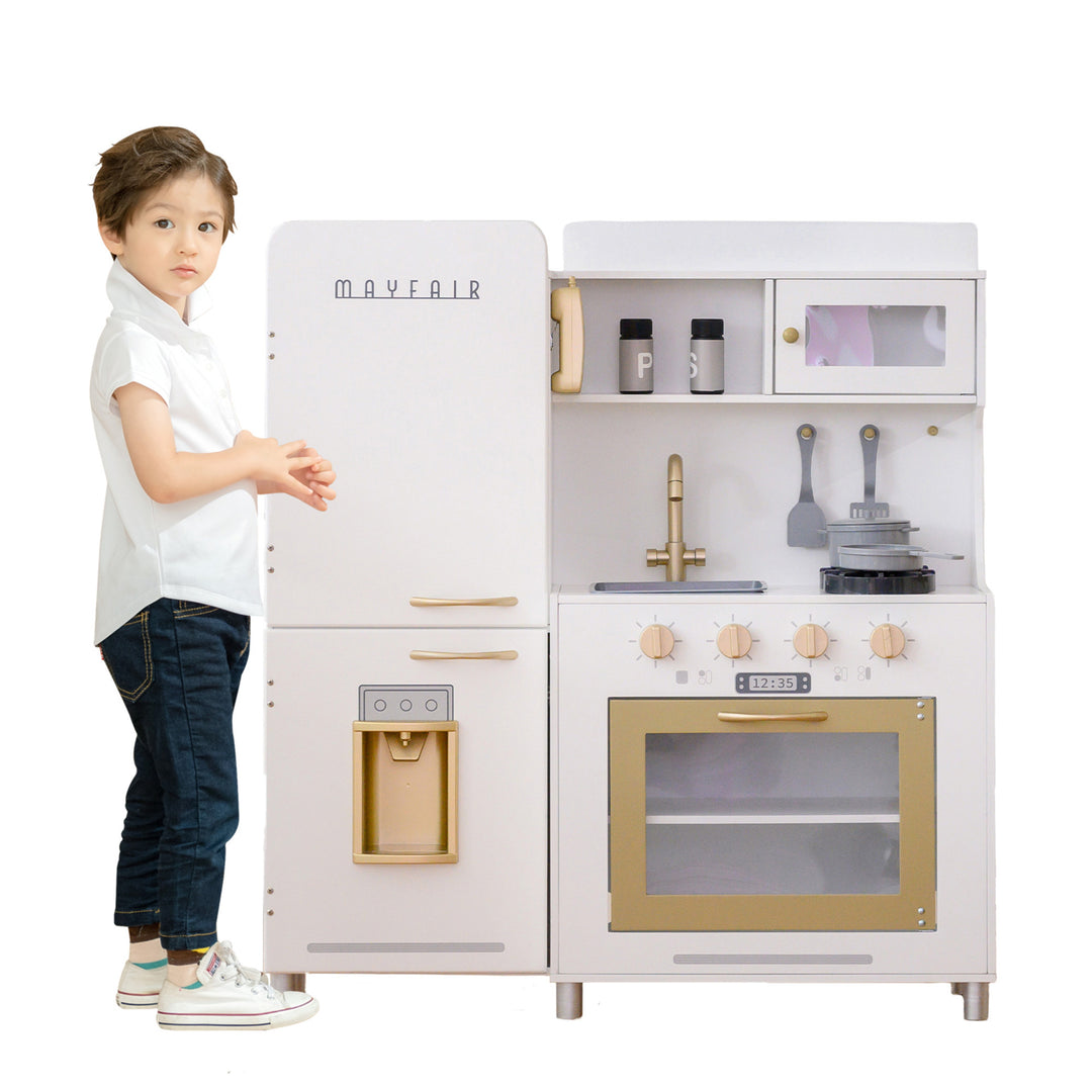 A child standing next to a Teamson Kids Little Chef Mayfair Classic Kids Kitchen Playset with 11 Accessories, White/Gold.