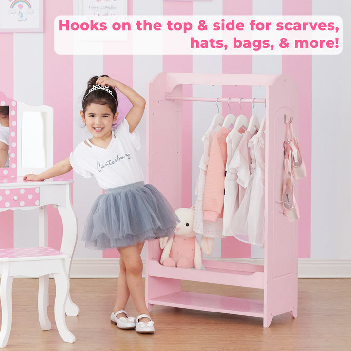 A little girl in a white tee and gray tutu standing in between a white and pink vanity and a pink wardrobe with dresses hanging from it in front of a white and pink striped wall.