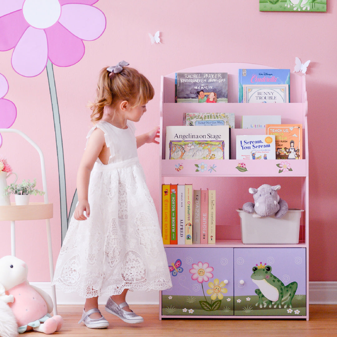 A little girl standing in front of a Fantasy Fields Magic Garden Kids 3-Tier Wooden Bookshelf with Storage, Multicolor.