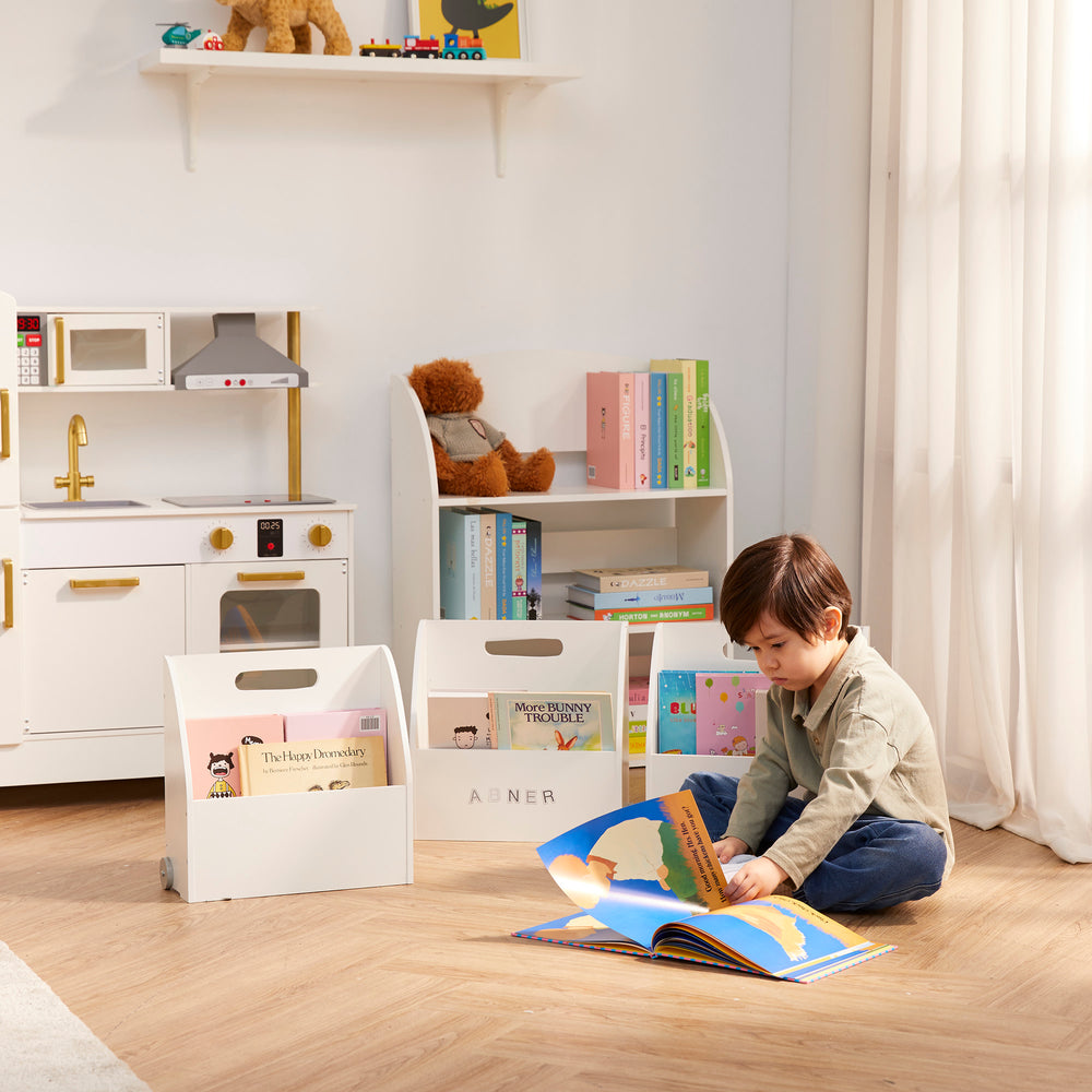 A little boy sitting on the ground reading a book next to hisFantasy Fields Kids Portable Bookcase Set of 3 with Magnetic Dry Erase Whiteboard, White